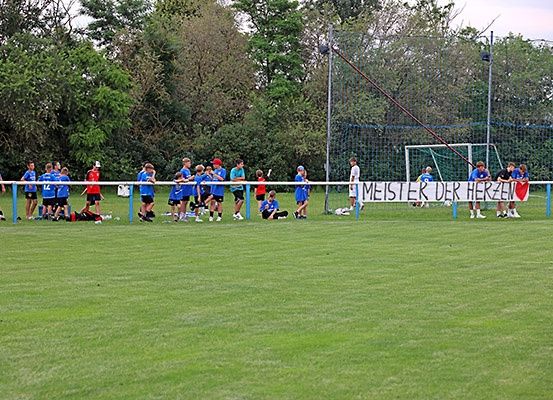 Eine Gruppe von Fußballspielern in blauen Trikots steht auf einem Feld, mit einem Banner, das 'MEISTER DER HERZEN' im Hintergrund liest.
