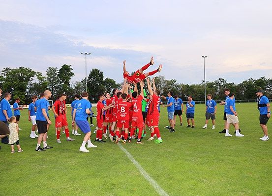 Eine Fußballmannschaft in roten Trikots feiert auf einem Feld, wobei Spieler einen Teamkollegen in die Luft heben, während andere und Zuschauer in Blau in der Nähe stehen, unter einem teilweise bewölkten Himmel.