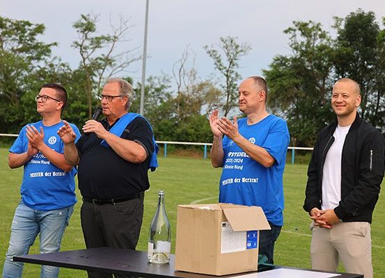 Vier Männer stehen auf einem Sportplatz und klatschen. Eine Flasche und eine Kiste stehen auf einem Tisch vor ihnen. Der Mann auf der linken Seite trägt eine Brille und ein Armband. Der Mann in der Mitte hält ein Mikrofon. Der Mann auf der rechten Seite trägt eine Jacke.