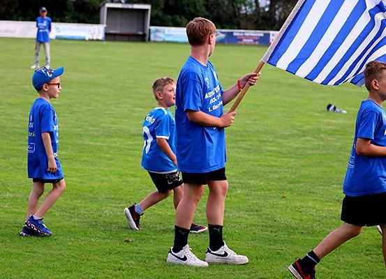 Mehrere Jungen in blauen Trikots gehen auf einem Grasfeld. Ein Junge hält eine Flagge. Dahinter steht eine Person auf dem Feld.