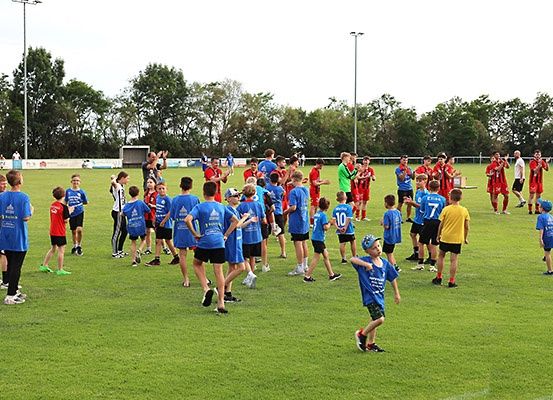 Eine Gruppe von Kindern und Erwachsenen in blauen und roten Trikots steht auf einem Feld, einige winken und andere beobachten, mit Bäumen und Straßenlaternen im Hintergrund.