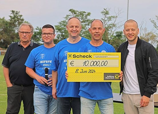 Fünf Männer stehen auf einem Feld, einer hält einen gelben Scheck über 10.000 Euro. Sie sind lässig gekleidet, zwei in blauen T-Shirts und drei in Freizeitkleidung. Der Mann in der Mitte hält den Scheck und lächelt. Dahinter ist ein unscharfer Hintergrund mit Bäumen und einem Tisch.