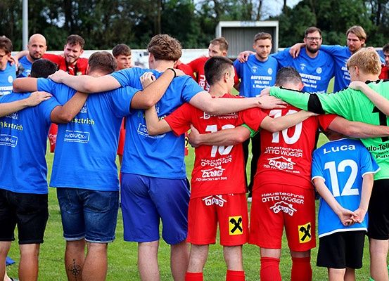 Eine Gruppe von Männern in Sporttrikots steht auf einem Rasenfeld eng beieinander, wahrscheinlich in Vorbereitung auf ein Spiel. Einige tragen Blau, andere Rot. Hinter ihnen ist ein unscharfer Hintergrund mit Bäumen und einem Gebäude.