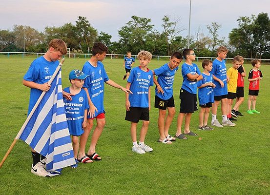 Gruppe junger Jungen in blauen Trikots, einige mit Mützen, aufgereiht auf einem Rasenfeld. Ein Junge hält eine blau-weiße Flagge.
