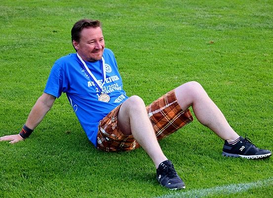 Ein Mann sitzt auf einem Rasenfeld und trägt ein blaues Shirt mit einer Medaille und braunen karierten Shorts. Er lächelt und lehnt sich auf seinen linken Arm.