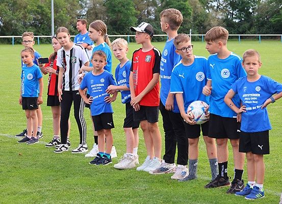 Eine Gruppe junger Fußballer in blauen Trikots steht auf einem Feld, einige halten einen Fußball. Dahinter sind Erwachsene und andere Kinder anwesend, die möglicherweise an einem Fußballevent teilnehmen.