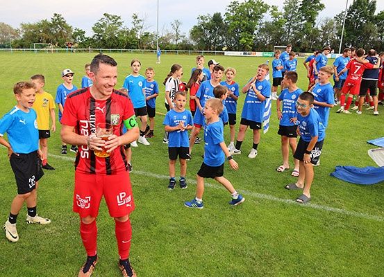 Ein Mann in Rot steht auf einem Fußballfeld mit einer Gruppe jubelnder Kinder. Sie tragen alle blaue Uniformen. Einige Kinder tragen Brillen, und eines hält einen Becher. Das Feld ist von Bäumen und einem Zaun umgeben.