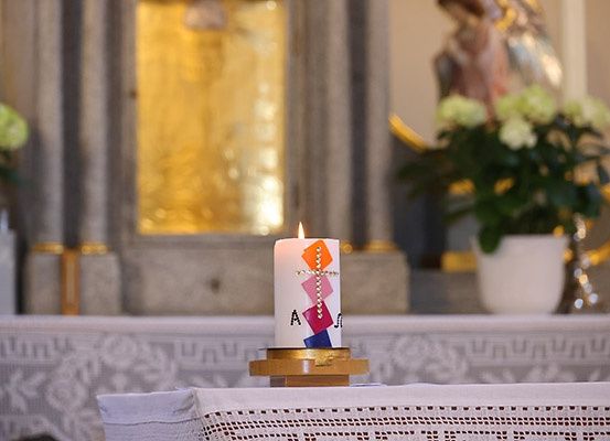 Eine brennende Kerze mit Kreuz und bunten Quadraten steht auf einem Tisch in einer Kirche. Sie befindet sich vor einem goldgerahmten Spiegel und einer Statue. Blumen stehen auf beiden Seiten des Spiegels.