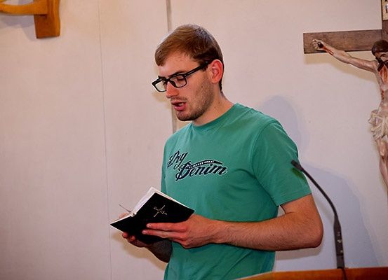 Ein Mann in einem grünen T-Shirt liest aus einem Buch, während er an einem Podium in einer Kirche steht. Hinter ihm ist ein Kruzifix an der Wand befestigt.