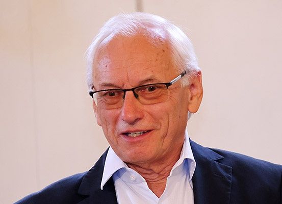 Ein älterer Mann mit weißen Haaren, der eine Brille und einen blauen Anzug trägt, lächelt in die Kamera.