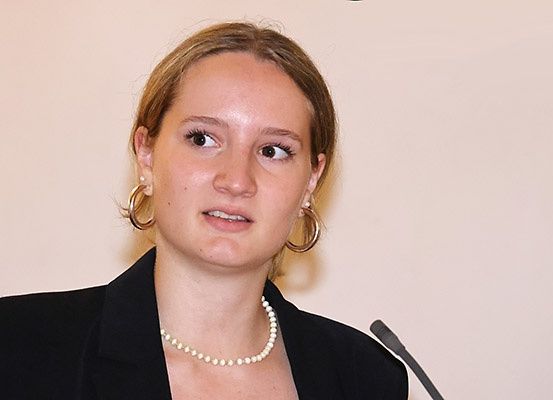Eine Frau mit kurzen blonden Haaren spricht in ein Mikrofon. Sie trägt einen schwarzen Blazer und eine Perlenkette.
