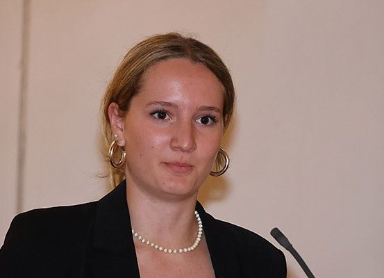Eine Frau in einem schwarzen Blazer, mit Perlenkette und Creolen steht vor einem Mikrofon und hält wahrscheinlich eine Rede.