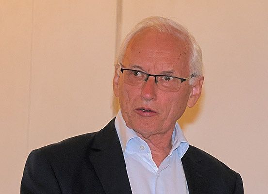Ein älterer Mann mit Brille und einem schwarzen Anzug steht vor einer weißen Wand. Er schaut nach rechts.