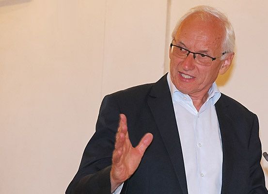 Ein älterer Mann in einem schwarzen Anzug und mit Brille steht an einem Rednerpult, gestikuliert mit seiner Hand und lächelt für ein Foto.