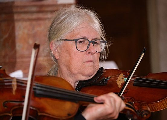 Eine ältere Frau mit grauen Haaren spielt eine Geige und trägt eine Brille. Sie hält einen Geigenbogen in der linken Hand.