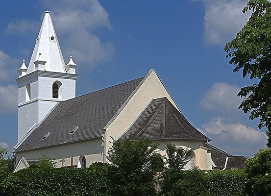 Eine weiße Kirche mit Turm und grauem Dach, umgeben von Bäumen und Büschen unter einem blauen Himmel mit Wolken.