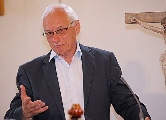 Ein älterer Mann mit Brille und dunklem Anzug steht an einem Rednerpult und gestikuliert mit der Hand. Ein Mikrofon steht vor ihm und ein Kruzifix ist an der Wand hinter ihm zu sehen.