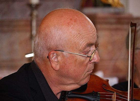 Ein älterer Mann mit Brille spielt Geige in einem schwach beleuchteten Raum. Er schaut nach unten, während er die Geige hält. Der Hintergrund ist unscharf.