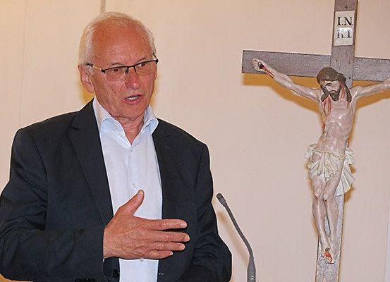 Ein älterer Mann in Anzug und Brille spricht an einem Podium mit einem Kruzifix im Hintergrund.