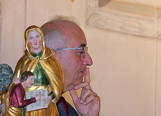 Ein älterer Mann mit Brille und Anzug steht vor einer Statue einer Frau mit einem Kind und berührt sein Kinn.