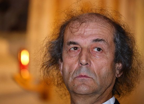 Nahaufnahme eines Mannes mit lockigem Haar und ernster Miene. Der Hintergrund ist unscharf, eine Lichtquelle beleuchtet von links.