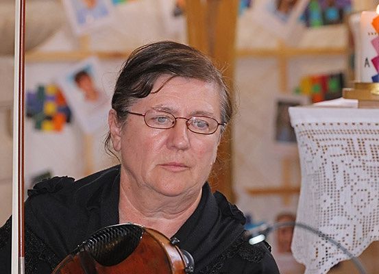 Eine Frau mit Brille spielt Geige in einem Raum mit Kerze und Bildern an der Wand.