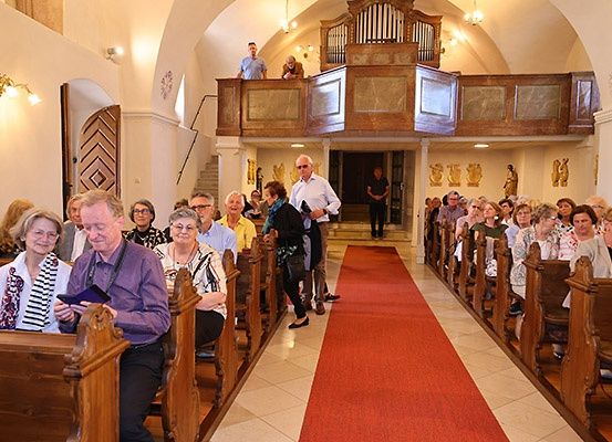 Eine Gruppe von Menschen sitzt in Kirchenbänken, einige stehen beim Eingang. Ein roter Teppich führt zur Vorderseite, und einige Personen stehen auf der Empore.