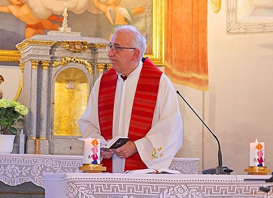 Ein Priester steht hinter einem Podium in einer Kirche, liest aus einem Buch, mit Kerzen und einem Mikrofon auf dem Tisch. Hinter ihm zeigt eine Wandmalerei Engel.