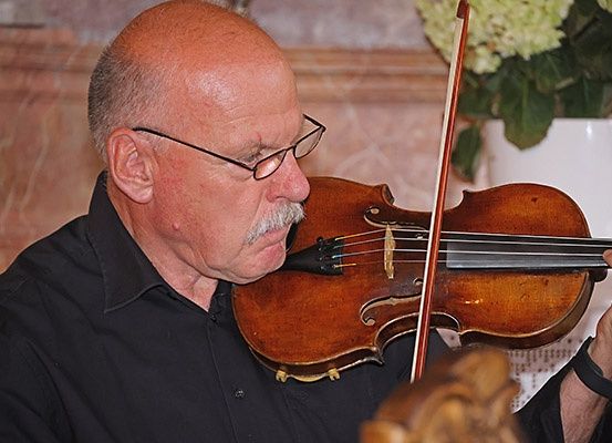 Ein älterer Mann mit Brille und einem Schnurrbart spielt eine Geige in einem Raum mit einer Blumenarrangement hinter ihm.
