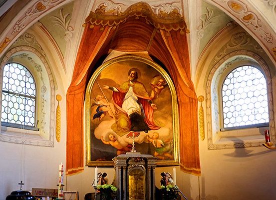 Eine Kircheninnenansicht mit einem großen gerahmten Gemälde eines Heiligen, umgeben von Engeln, positioniert über einem kleinen Altar mit Kerzen.