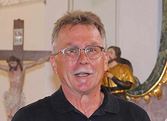 Ein älterer Mann mit Brille und Schnurrbart lächelt und steht vor einem Kruzifix und einem Spiegel.