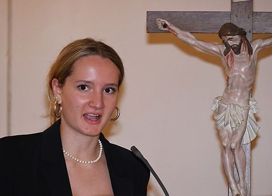 Eine Frau mit blonden Haaren, die eine Perlenkette und Ohrringe trägt, spricht in ein Mikrofon, mit einem Kruzifix hinter ihr.