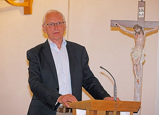 Ein älterer Mann mit Brille und Anzug steht an einem Podium vor einem Kreuz mit einer gekreuzigten Figur.