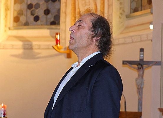 Ein Mann in einem Anzug singt in einer Kirche mit Kerzen und einem Kreuz an der Wand hinter ihm.