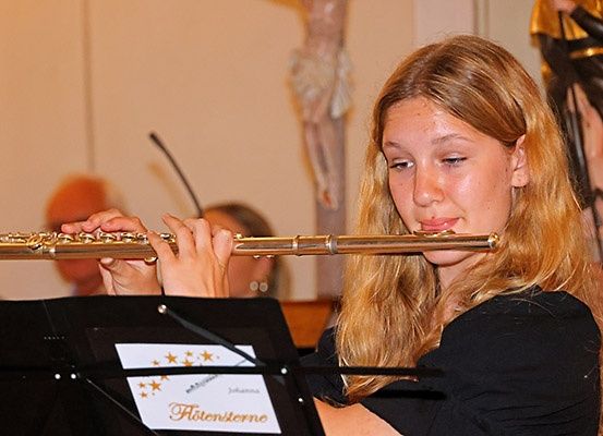 Eine junge Frau spielt eine Flöte während einer Aufführung in einer Kirche. Sie trägt ein schwarzes Oberteil und hat blonde Haare. Ein Notenständer mit Namenskarte steht vor ihr.