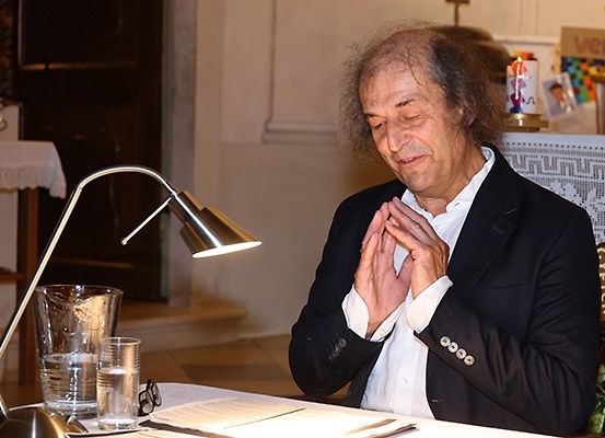 Ein älterer Mann mit grauem Haar betet in einer Kirche, er trägt einen schwarzen Anzug. Er sitzt an einem Tisch mit einer Lampe, Wassergläsern und einem Buch. Hinter ihm steht ein Schreibtisch mit einem Kreuz und Kerzen.