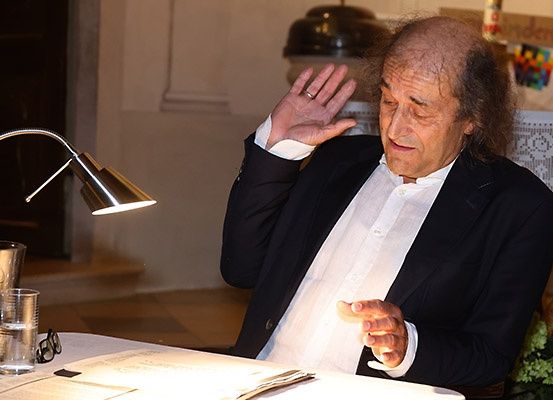 Ein älterer Mann mit einem Ring an der Hand sitzt an einem Tisch mit einer Lampe und einem Glas Wasser auf dem Tisch.