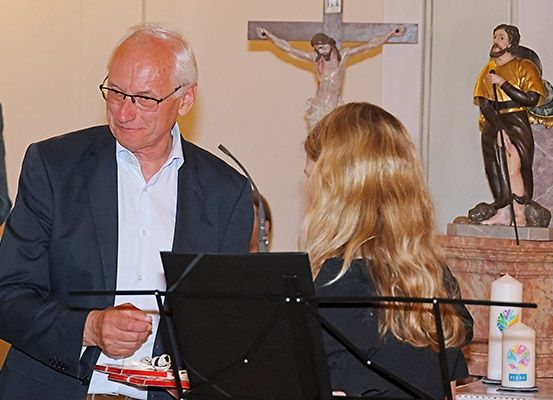 Ein älterer Mann und eine Frau stehen vor einem Notenständer in einer Kirche, der Mann hält ein kleines Objekt und ein Mikrofon, die Frau schaut ihn an, mit einem Kreuz und Statuen hinter ihnen.