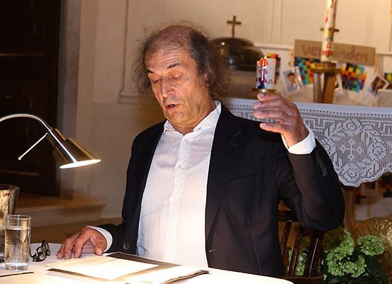 Ein älterer Mann mit langen Haaren sitzt an einem Tisch mit einem Glas Wasser und einem Buch, hält eine Kerze und liest einen Text. Hinter ihm steht eine Lampe, eine weiße Tischdecke und eine Kerze auf einem Ständer.