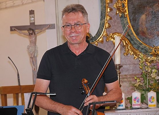 Ein Mann mit Brille und schwarzem Poloshirt hält eine Geige vor einem Altar mit Kreuz und Kerzen.