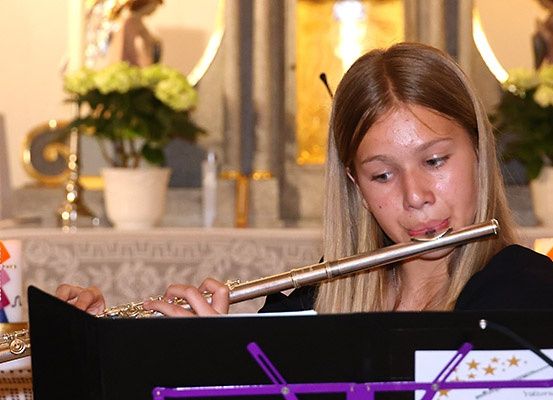 Ein junges Mädchen spielt eine Flöte in einer Kirche, mit Blumen und Kerzen im Hintergrund.