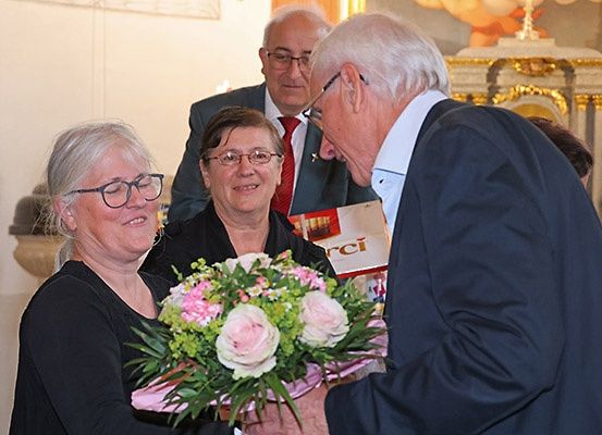 Zwei Frauen halten einen Blumenstrauß, während ein Mann mit Brille eine von ihnen auf die Wange küsst. Ein anderer Mann hinter ihnen trägt einen Anzug und eine Krawatte. Der Hintergrund zeigt einen Kirchenaltar.
