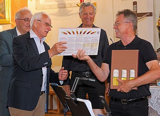 Fünf Männer in Anzügen und Hemden stehen in einer Kirche, einer hält ein Plakat und ein anderer eine Schachtel. Die Männer lächeln und hinter ihnen stehen Statuen.