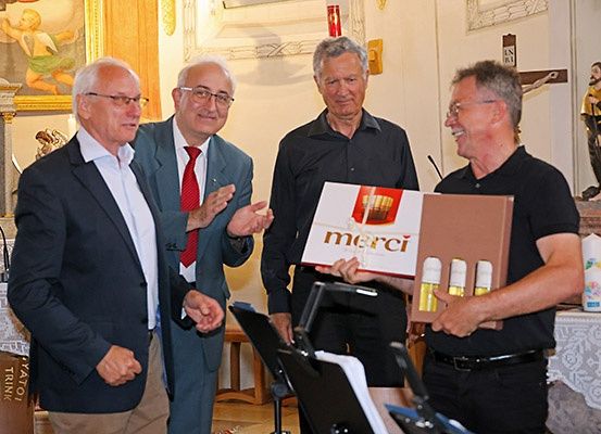 Vier Männer in einer Kirche mit einem Podium, Notenblättern und einer 'merci'-Box. Zwei Männer in Anzügen, einer mit roter Krawatte, einer in Schwarz. Zwei lächelnde Männer, einer hält die Box.