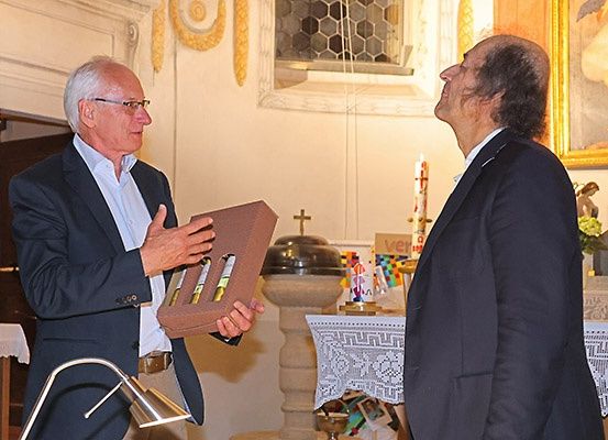 Zwei Männer in Anzügen stehen in einer Kirche, einer hält eine Box und spricht, der andere schaut nach oben. Hinter ihnen steht ein Tisch mit einem Kreuz und Kerzen.