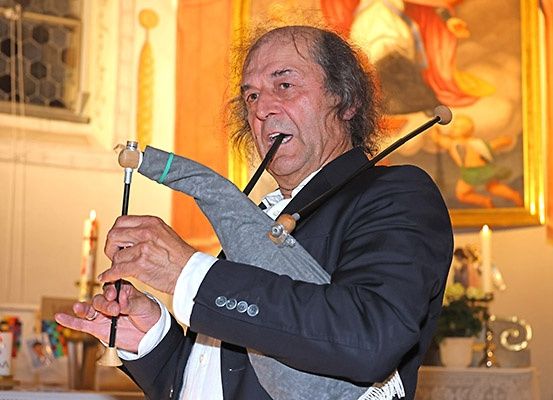 Ein älterer Mann spielt Dudelsack und hält zwei Pfeifen in einer gerahmten goldenen Umgebung mit Kerzen und einem Tisch mit Blumen.