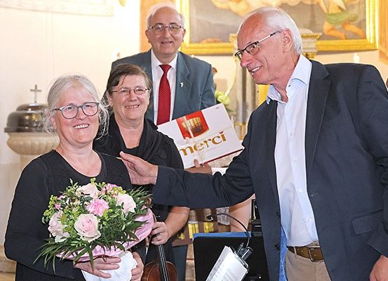 Ein Mann, der ein Buch mit der Aufschrift 'merci' hält, überreicht einer Frau in einem schwarzen Shirt Blumen. Dahinter hält eine weitere Frau ein Cello und ein Mann mit roter Krawatte lächelt.