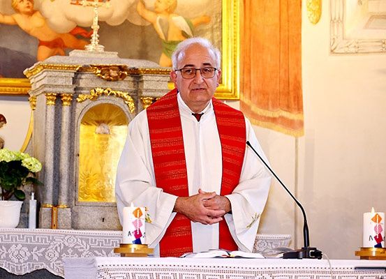 Ein Priester in einer roten Stola steht an einer Kanzel in einer Kirche, mit Kerzen und Blumen im Hintergrund.