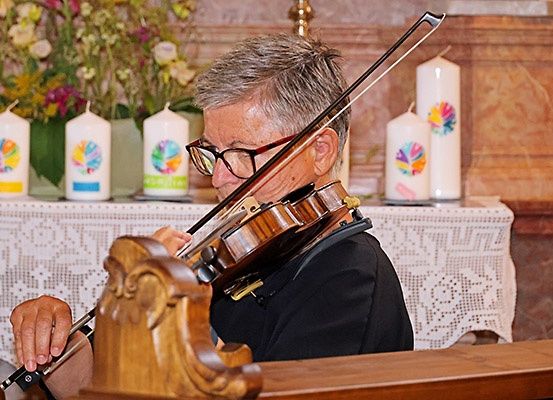Ein älterer Mann spielt eine Geige in einer Kirche, umgeben von Kerzen und Blumen.