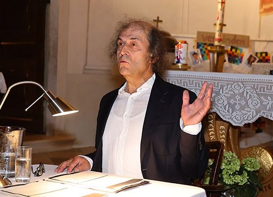 Ein älterer Mann sitzt an einem Tisch und gestikuliert mit der Hand. Er trägt einen schwarzen Anzug und ein weißes Hemd. Hinter ihm befindet sich ein weißer Altar, geschmückt mit einer Kerze und einem Schild mit der Aufschrift 'Verbinden'. Ein Mikrofon und eine Lampe stehen auf dem Tisch.